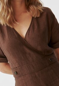 Braunes Leinenwickelkleid mit kurzen Ärmeln, V-Ausschnitt und dekorativen Knöpfen an der Taille. Die Textur wirkt leicht und leicht zerknittert.