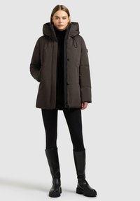 Giacca parka nera imbottita con cappuccio, caratterizzata da una cerniera frontale, bottoni a scatto e tasche laterali. Indossata sopra un dolcevita nero e leggings.