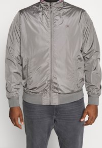 Man wearing a zipped gray satin bomber jacket with ribbed cuffs and hem, paired with dark gray jeans, hands relaxed at sides.