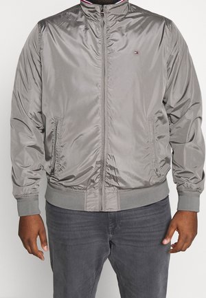 Mand iført en grå satin bomberjakke med lynlås, ribbede manchetter og nederste kant, sammen med mørkegrå jeans, med hænderne afslappet ved siden af.