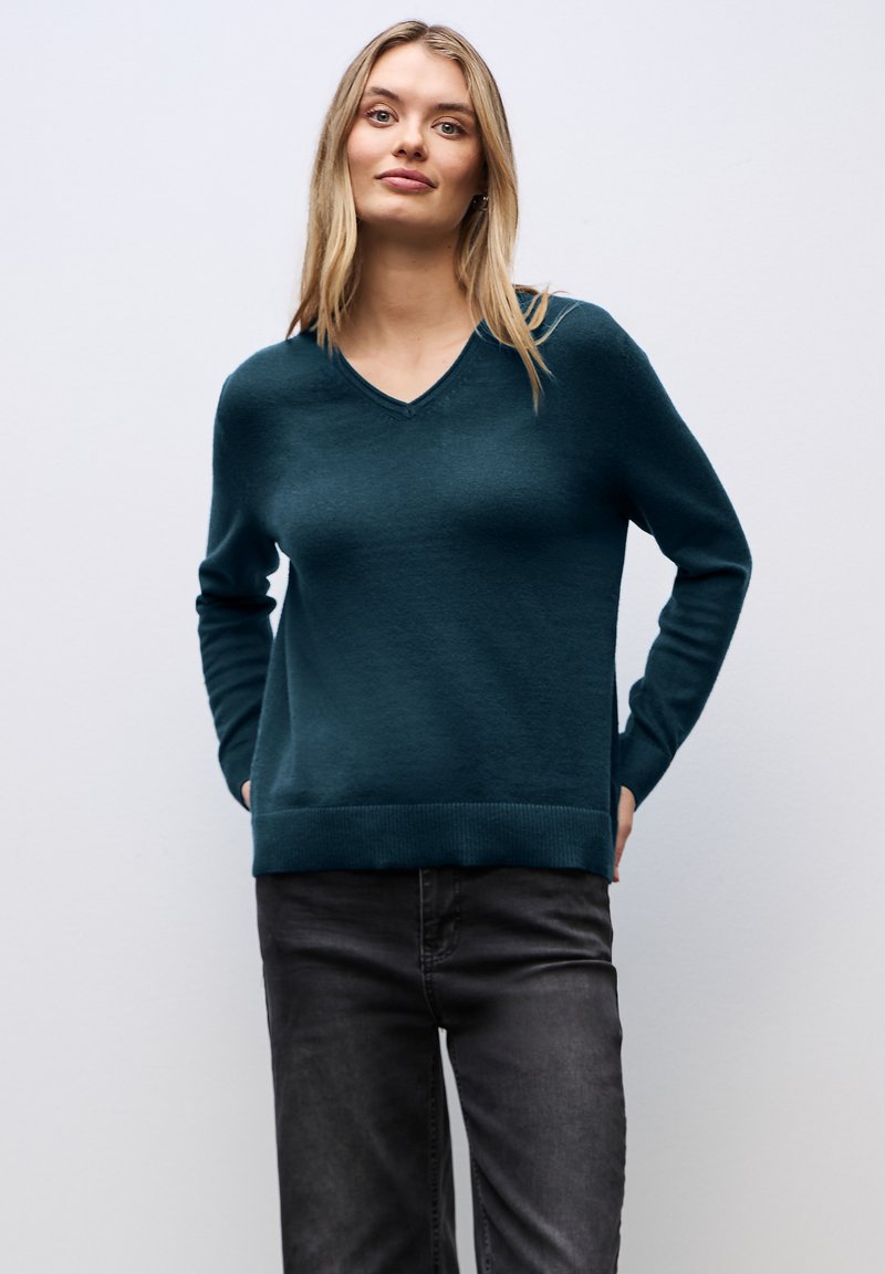 Teal V-ringad tröja med långa ärmar, ribbad nederkant och slät textur, i kombination med mörka jeans. Minimalistisk design mot en neutral bakgrund.