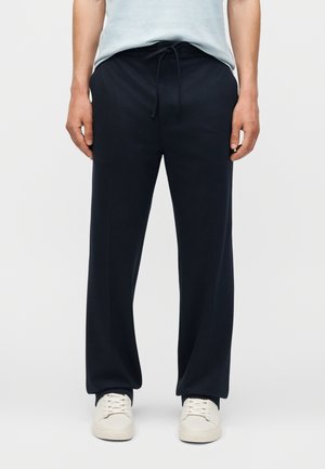 Homme portant un pantalon à cordon bleu marine et des baskets blanches, debout devant un fond clair uni, les mains détendues sur les côtés.