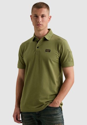 Homme portant un polo vert olive à manches courtes avec col et poche, associé à un jean bleu, debout devant un fond uni.