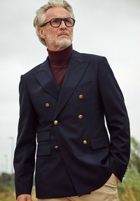 Marineblauwe dubbelgeknoopte blazer met gouden knopen, gecombineerd met een bordeauxrode coltrui. Gestructureerde stof, op maat gemaakt pasvorm en opvallende revers.