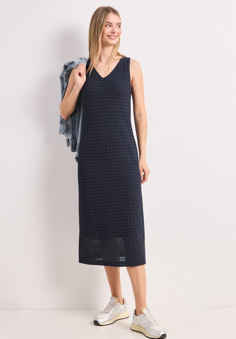 Femme portant une robe midi texturée bleu marine sans manches avec des baskets blanches, tenant une veste en jean sur son épaule, debout contre un mur blanc.