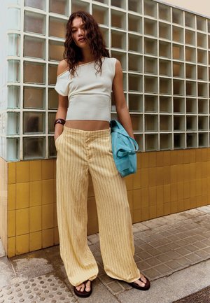 Woman with curly hair wearing cream one-shoulder top, loose yellow striped pants, black sandals, holding blue bag by tiled wall.