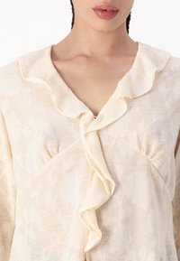 Helle beigefarbene Bluse mit einem Rüschenausschnitt, versehen mit floralen Mustern und einem strukturierten, halbtransparenten Stoff. Verschluss mit kleinen metallischen Details.