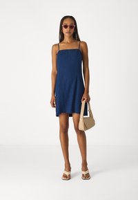 Robe sans manches bleu avec un décolleté carré et une coupe ample, assortie à un sac à main beige et des sandales de couleur claire.