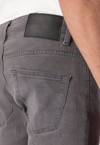 Jeans in denim grigio con una finitura testurizzata, tasche posteriori e un'etichetta in pelle nera impressa con il nome del marchio.