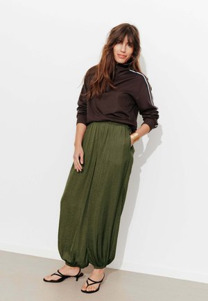 Femme aux longs cheveux bruns portant un haut noir à fermeture éclair avec des rayures blanches et un pantalon ample vert olive avec des sandales noires à brides.