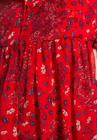 Rote Bluse mit Blumenmuster, die ein plissiertes Design, eine Knopfleiste und kleine lila und weiße Blumenmuster auf leichtem, strukturiertem Stoff aufweist.