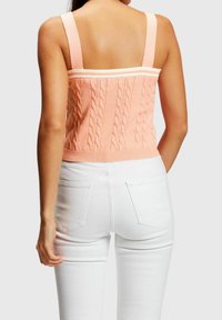 Brzoskwiniowy crop top z dzianiny warkoczowej z szerokimi ramiączkami, wyposażony w kontrastujący biały prążkowany pas z tyłu. Noszony z białymi dopasowanymi spodniami.