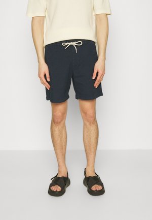 Shorts en coton bleu marine avec un waistband élastique et un cordon de serrage. Portés avec des sandales noires, debout contre un fond blanc uni.