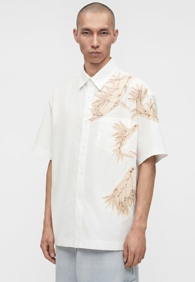 Άνδρας που φοράει λευκό κοντομάνικο πουκάμισο με μπεζ αφηρημένα βοτανικά σχέδια στη δεξιά πλευρά και ανοιχτό μπλε παντελόνι.