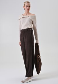 Maglione color crema con spalle scoperte, pantaloni marroni a vita alta e sneakers beige. La modella tiene una borsa marrone scuro con texture.