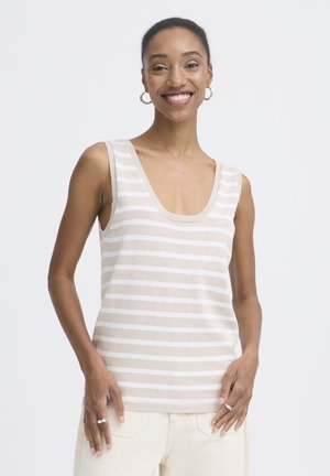 Mujer sonriente con top sin mangas de rayas beige y blancas y pantalones de color claro, con pendientes de aro y anillos en ambas manos.