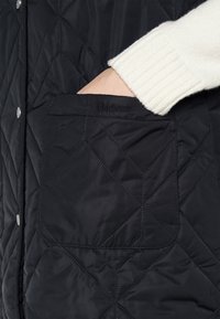 Schwarze gesteppte Weste mit einer strukturierten Tasche. Verfügt über silberne Druckknöpfe und ein besticktes "Barbour"-Logo. Darunter sichtbar ein weißer Rippstrickpullover.