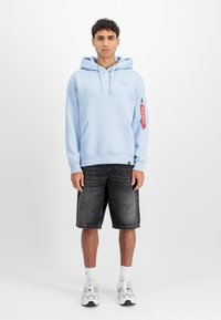 Világoskék kapucnis pulóver elülső zsebbel és cipzáras ujjzsebbel, sötét farmer rövidnadrággal és futócipővel párosítva.