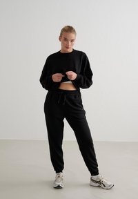 Schwarze, kurze Sweatshirt mit langen Ärmeln und besticktem Detail, kombiniert mit schwarzen Jogginghosen mit elastischem Bund. Sportliche Schuhe.