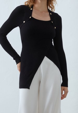 Femme portant un haut noir côtelé à manches longues avec des boutons dorés sur un pantalon blanc à jambes larges, sur fond uni.