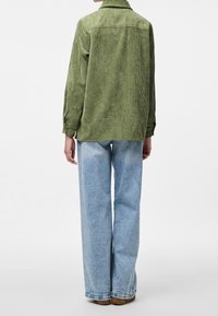 Zielona sztruksowa koszula z kołnierzykiem, długimi rękawami i prostym tyłem. W połączeniu z jasno-niebieskimi szerokimi dżinsami i brązowymi butami.