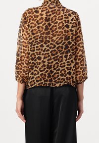 Blouse transparente à imprimé animal avec un motif léopard marron et beige, manches trois-quarts et coupe ample. Associée à un pantalon noir.
