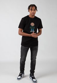 Černé tričko s grafickým designem postavy v zeleném kabátu, spárované se šedými džíny a modrými vysokými teniskami.