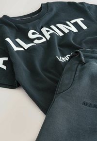 Tume roheline puuvillane t-särk, millel on suur valge tekst "ALLSAINT" ja detail "LONDON". Koos sobivate tekstuuriga dressipükstega.