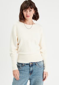 Cremefarvet strikket sweater med rund halsudskæring, ribbede manchetter og kant. Båret med højtaljede blå denim jeans.