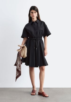 Žena nosiace čierne krátke šaty so sťahovacou šnúrkou v páse, drží béžovú pletenú tašku a šál s paisley vzorom, nosí červené šľapky.