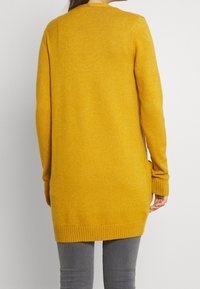 Gul stickad cardigan, lång med ribbade ärmslut och nederkant, med mjuk textur och sidofickor, visad från baksidan.