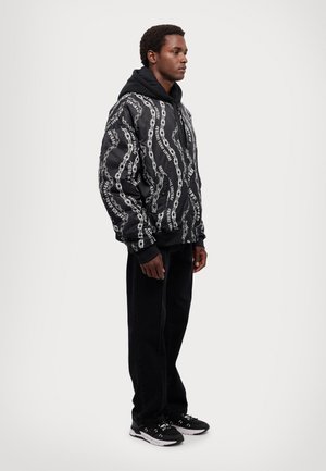 Veste bombardier noire avec un motif de chaîne blanche, dotée d'une capuche et de poignets côtelés, associée à un pantalon droit noir et des baskets noires.