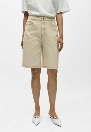 Beige knelange denimshorts med frontlommer, brukt med hvit topp og spisse hvite slip-on sko på hvit bakgrunn.
