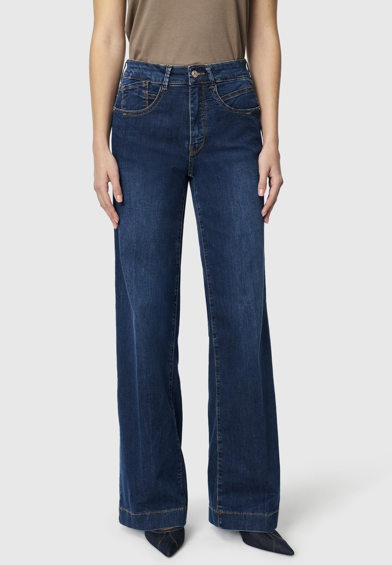 Persona con jeans vaqueros de pierna ancha de tiro alto en azul oscuro, camiseta sin mangas marrón metida por dentro y zapatos negros puntiagudos.