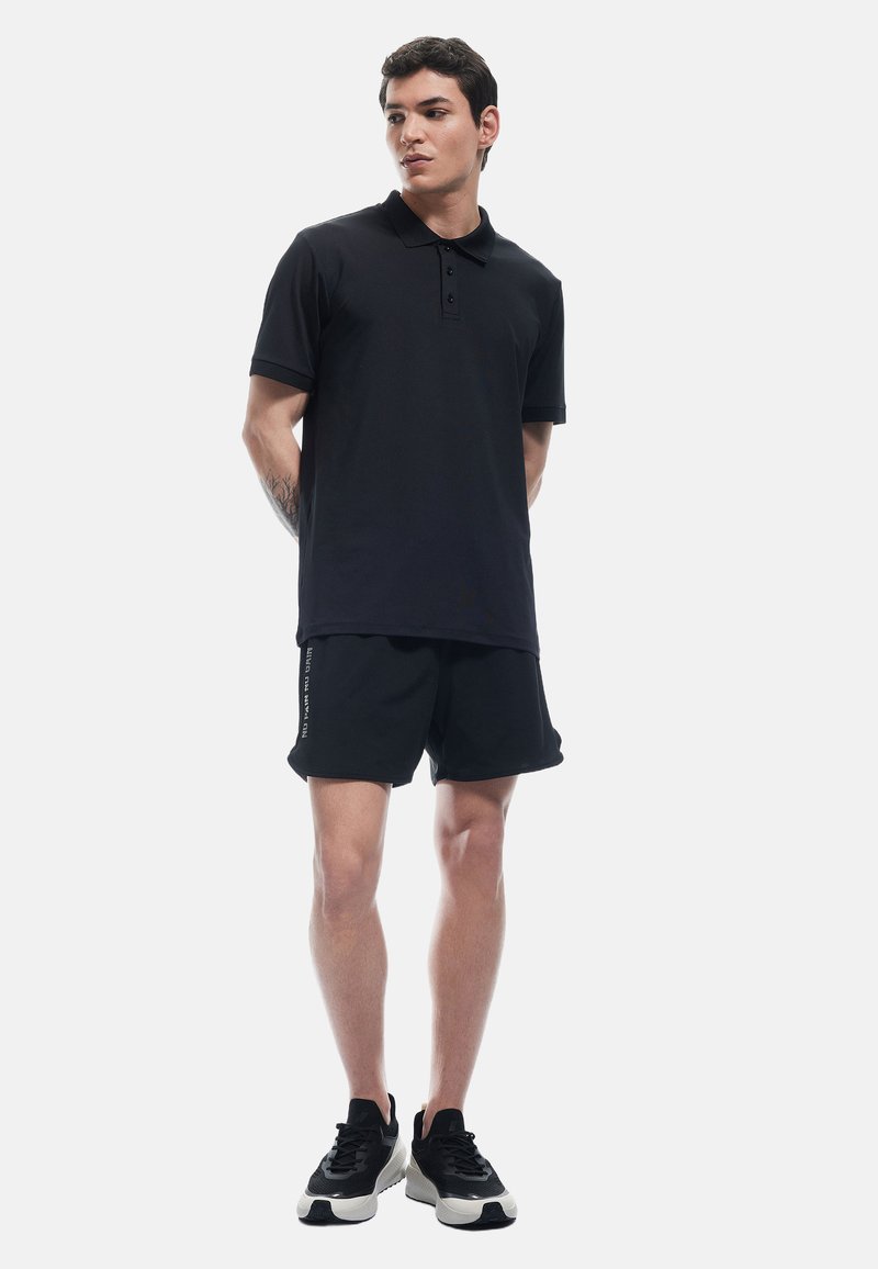 Sort polo-shirt med en tre-knaps lukning, parret med sorte shorts med et side-logo print. Båret med sort og hvidt sneakers.