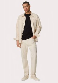 Hellbeiges Hemd mit Knöpfen über einem schwarzen T-Shirt, kombiniert mit weißen Hosen und hellen Sneakers; moderner Schnitt, glatte Textur, minimalistische Details.