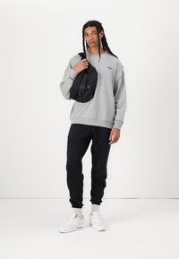 Sweatshirt cinza com logótipo azul, mochila preta, calças de esporte pretas e ténis brancos. Textura suave, ajuste descontraído e punhos ajustados.