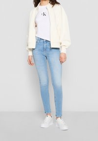 Vit zip-up bomberjacka med logotyp på bröstet, kombinerad med ljusblå skinny jeans och vita sneakers. Slätt tyg och modern passform.