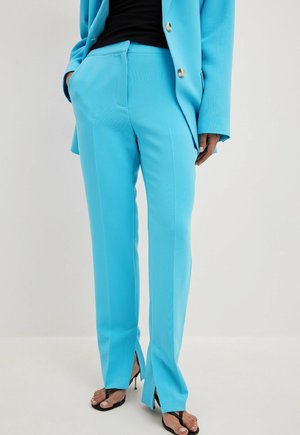 Frau trägt hellblaue, maßgeschneiderte Hosen mit vorderen Schlitzen, kombiniert mit einem passenden Blazer, schwarzem Top und schwarzen Riemchen-Absätzen.