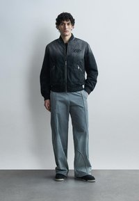 Vyras stovi dėvėdamas juodą bomber stiliaus striukę su užtrauktuku ir laisvas pilkas kelnes su matomais siūlais, juodus batus, prie paprasto šviesaus fono.