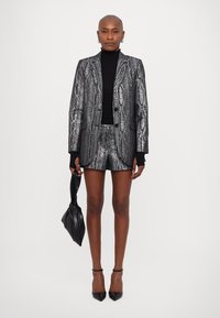 Metalický strieborný blazer a nohavicový set so vzorom hadej kože, skombinovaný s čiernym rolákom. Doplnky zahŕňajú čiernu kabelku.