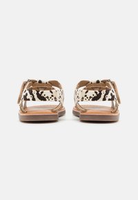 Pár plochých sandálů s černobílými tečkovanými pásky a hnědými podešvemi, pohled z zadní strany na bílém pozadí.