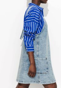 Robe salopette en denim bleu clair avec une grande poche avant, portée par-dessus une chemise à rayures bleues et blanches à manches longues avec des poignets boutonnés.