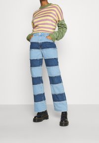 Suéter a rayas en verde, amarillo y púrpura; jeans patchwork en azul y azul claro con bordes deshilachados; botines negros gruesos.