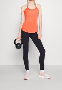 Helle orange ärmellose Sport-Tanktop, schwarze Leggings und hellfarbene Sneakers. Das Modell hält ein schwarzes Kettlebell.