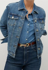Chaqueta de denim en azul claro con bolsillos con botones, un cuello clásico y detalles de metal plateado, combinada con una camisa de botones azul y un cinturón marrón.