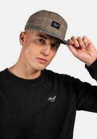 Braune Karokappe mit flachem Schild, versehen mit einem schwarzen Logo-Patch. Getragen mit einem schwarzen Sweatshirt, das weißen Text auf der Brust zeigt.