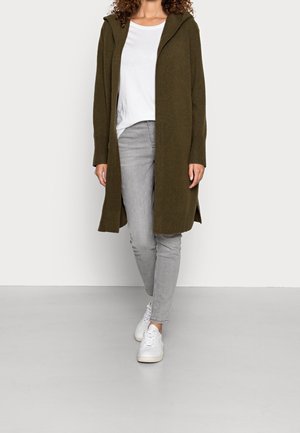 Woman wearing an olive green open-front long cardigan, white shirt, light gray jeans, and white sneakers, standing on a light gray floor.