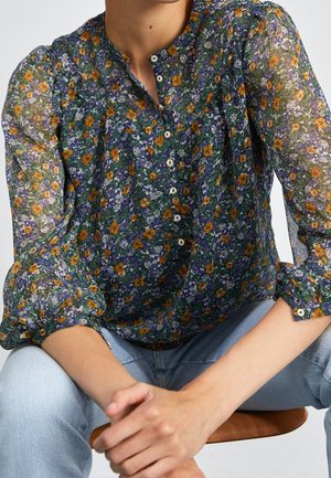 Nainen pukeutuneena ohueseen, pitkähihainen kukkakuosiseen puseroon, jossa on oransseja ja violetteja kukkia, napitettu edestä, yhdistettynä vaaleansinisiin farkkuihin, istuu kädet yhteen kiedottuina.