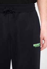 Sorte træningsbukser lavet af glat materiale med en elastisk talje. Har et grønt EA7-logo på venstre lår og en snøredetalje.